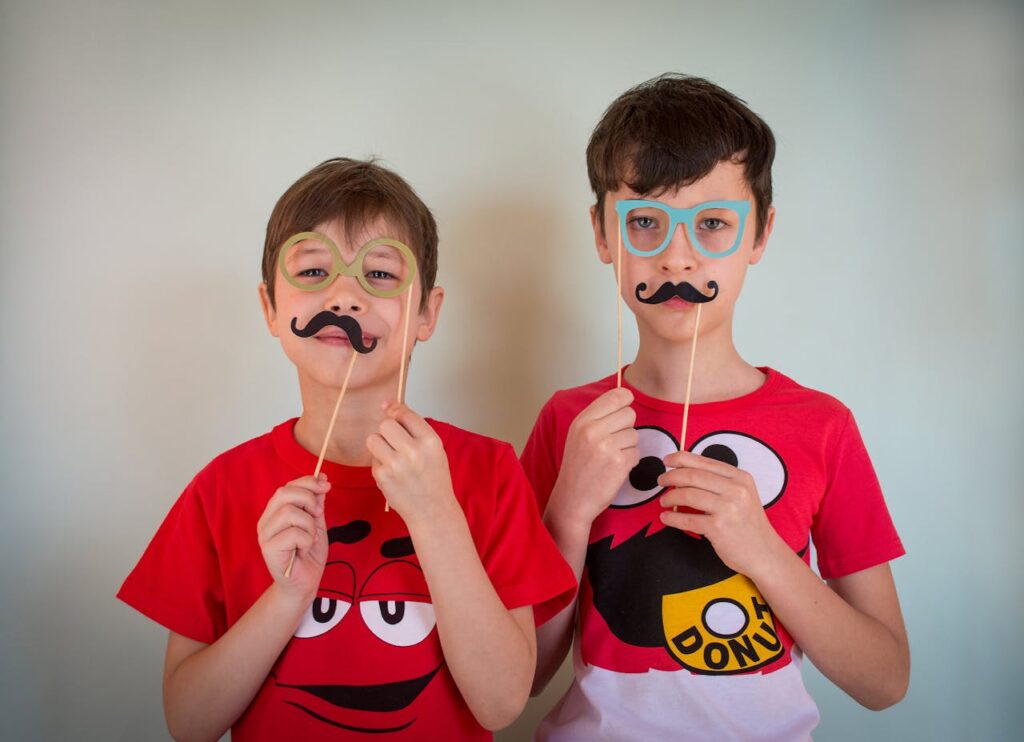 Happy two young males in party glasses and party mustache looking at camera while standing against white background